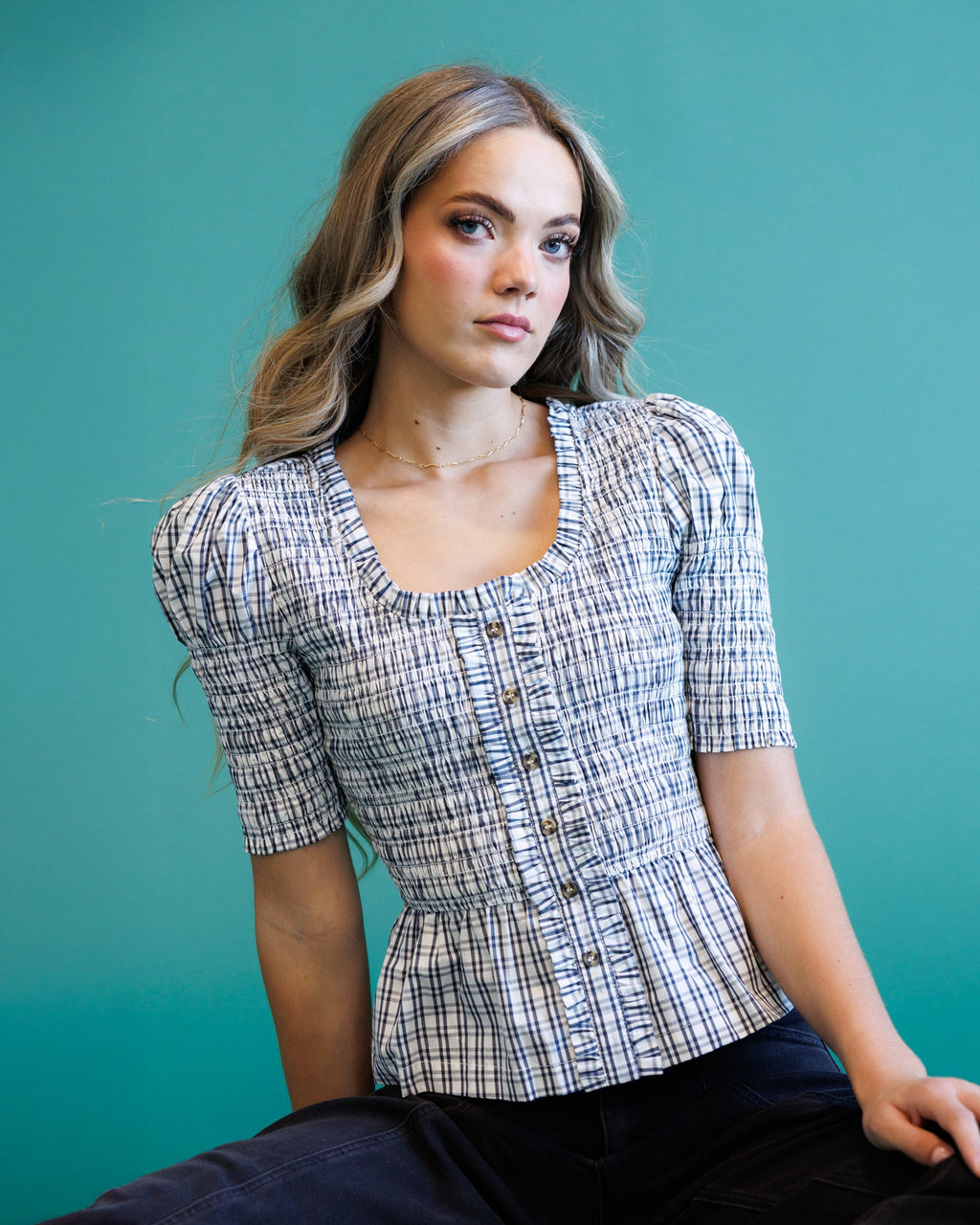 A woman wearing a short-sleeve plaid peplum top with puff sleeves and a buttoned front, posed against a solid teal background.