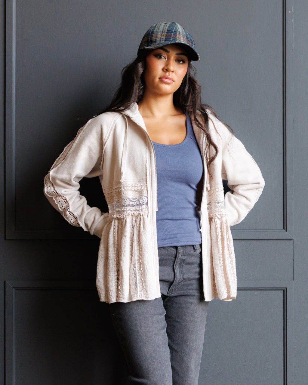 A woman wearing a cream-colored lace-trimmed hooded jacket over a blue tank top and gray jeans, standing against a dark gray paneled wall.