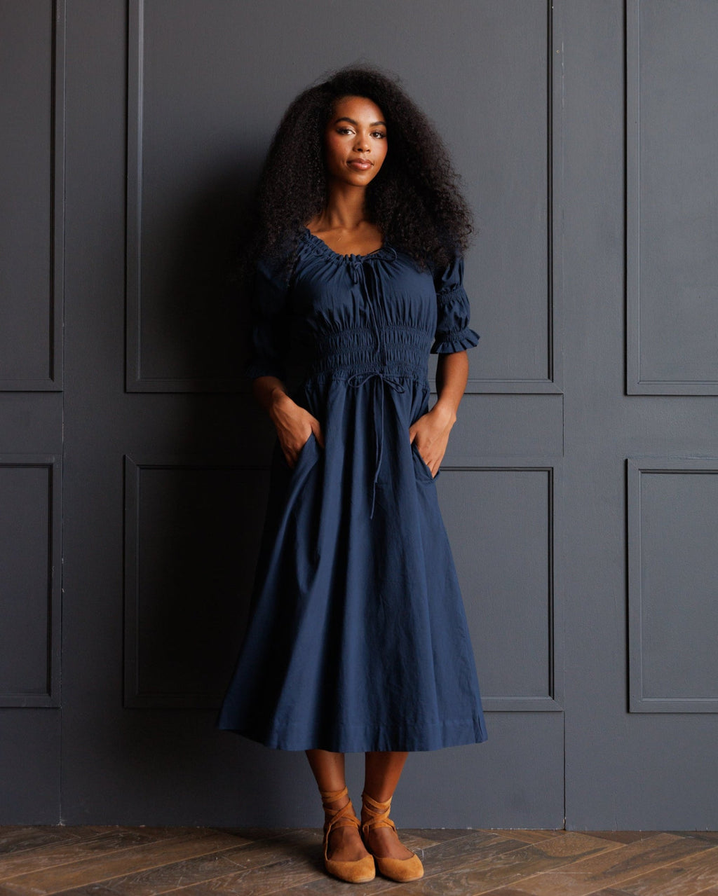 A woman with long, curly black hair stands against a dark gray paneled wall wearing a navy blue midi dress with puff sleeves, a smocked bodice, and side pockets. She is wearing tan ankle boots.