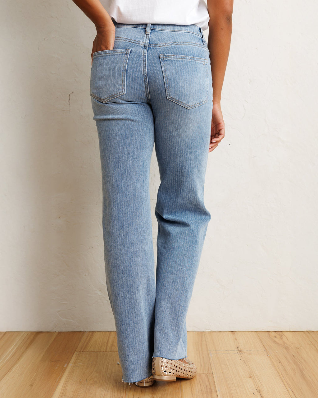 A person shown from the back wearing light-wash blue jeans, a white top, and beige perforated shoes, standing against a textured off-white wall on a light wood floor.