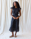 A woman modeling a dark navy midi dress with a delicate white and yellow floral print, featuring cap sleeves, a gathered waist, and a ruffled hem, standing against a draped white fabric backdrop.