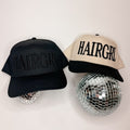 Two baseball caps displayed on mirrored disco ball props against a plain light background. The left cap is black with tonal black embroidery reading 'HAIRGRL'.